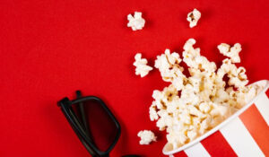 Noticias Cine muestra un recipiente de palomitas de maíz derramadas junto a unas gafas 3D sobre un fondo rojo, representando noticias cine y la experiencia cinematográfica. Esta imagen es adecuada para artículos sobre estrenos, críticas y festivales de cine.