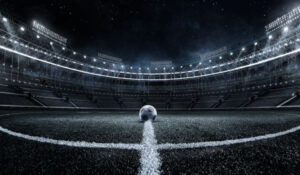 Estadio de fútbol iluminado de noche con una pelota en el centro del campo, representando Noticias Fútbol.