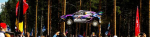 Coche de Rally en el aire durante un evento, rodeado de espectadores y banderas, ilustrando la historia de los Rallyes y la adrenalina de las carreras.