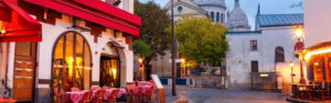 Calle en París, viaje a Francia, con un café al aire libre y la Basílica del Sagrado Corazón en el fondo al atardecer.