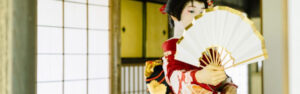 Geisha en kimono rojo con detalles dorados sosteniendo un abanico en un entorno japonés clásico, representando la rica cultura japonesa y los lugares increíbles para visitar como el parque Yoyogi.