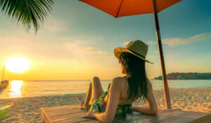 Mujer relajándose bajo una sombrilla en una playa tropical al atardecer, representando Noticias Playas.