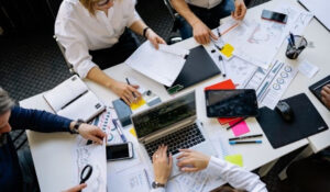 Noticias Trabajo muestra un grupo de personas trabajando en una mesa llena de documentos y dispositivos, representando noticias trabajo y la colaboración en el entorno laboral. Esta imagen es adecuada para artículos sobre ofertas de empleo, búsqueda de trabajo y trabajo remoto.